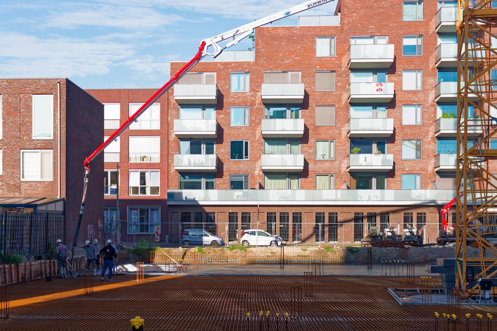 The red and white boom of a Schwing concrete pump arching over the Circulus rebar deck, its end hose lowered to a crew in white hard hats pouring the first concrete along the timber retaining wall, rust-orange rebar mesh with yellow-capped starter bars in the foreground and a brick apartment block with glass balconies behind
