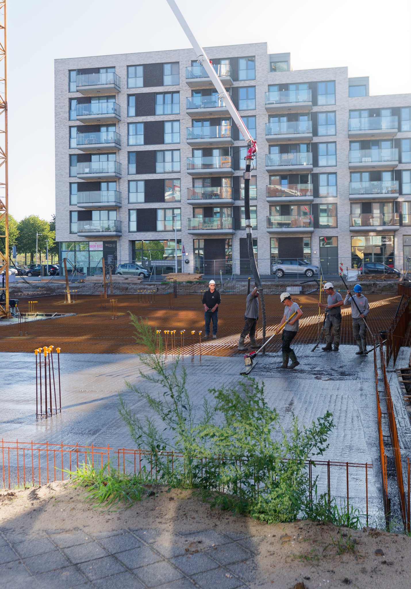 Crew pouring and levelling the Circulus foundation slab: one worker holding the black pump hose, another pulling a yellow vibrating screed through wet grey concrete, two more with long tools, a foreman in a black jacket standing on the uncovered rebar mesh ahead, pale brick apartment block with balconies behind and weeds and paving in the foreground